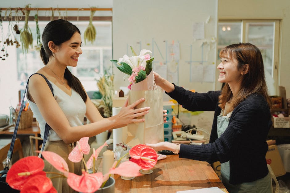 A young woman with dark hair tied back and a beige sleeveless top receives a floral gift from an older woman with brown hair, wearing a dark sweater and light-colored apron, inside a well-lit flower s