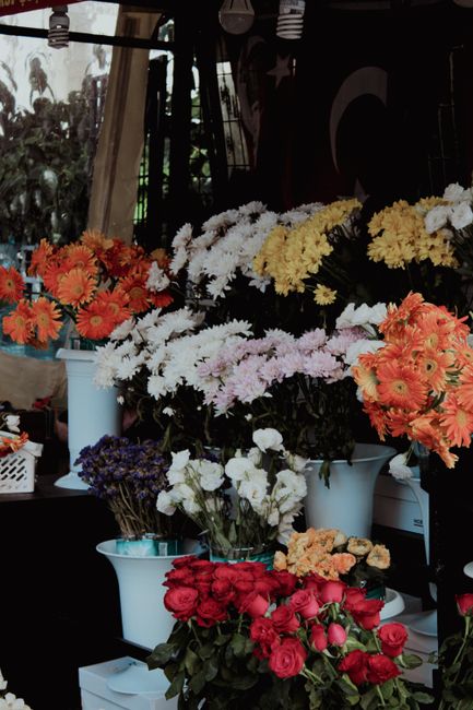 A display of various fresh flower arrangements at a floral shop, featuring vibrant orange gerbera daisies, soft white chrysanthemums, pale pink carnations, deep red roses, and purple statice. The flow