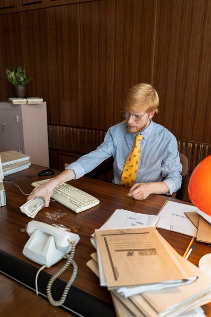 A man with reddish hair, glasses, and a beard, dressed in a light blue shirt and a yellow tie with black patterns, sitting at a wooden office desk. He is reaching across the desk to pick up a bundle o