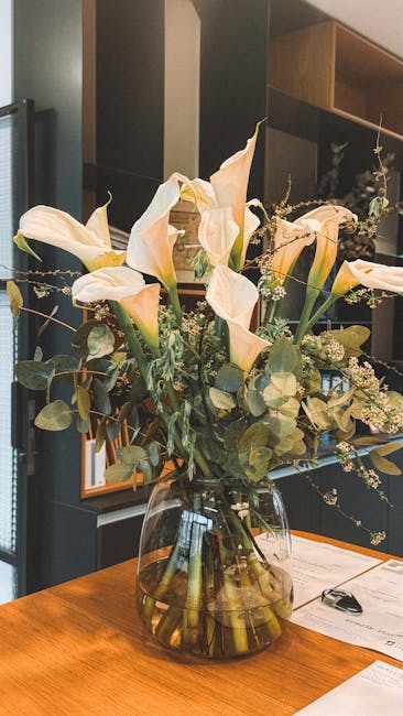 A tall, elegant floral arrangement featuring large, ivory calla lilies with smooth, trumpet-shaped petals, complemented by delicate sprigs of baby's breath and various eucalyptus leaves with rounded,