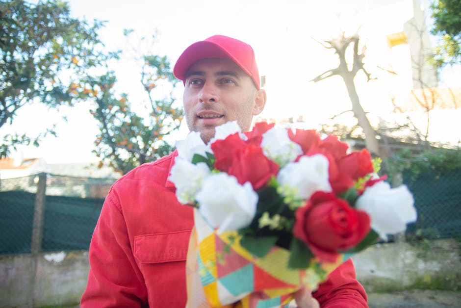 A male delivery person dressed in a red jacket and matching red cap holding a colorful bouquet of fresh flowers, including red and white roses, wrapped in yellow paper with a checkered pattern, outsid