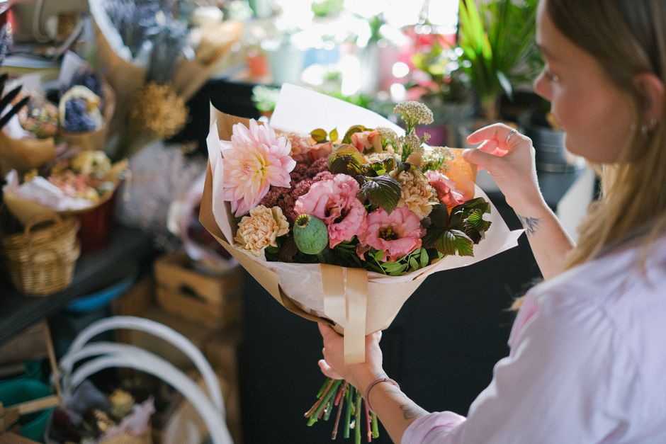 A woman with blonde hair, wearing a white shirt, is holding a bouquet of fresh flowers wrapped in beige paper and tied with a ribbon inside a flower shop. The bouquet includes large pink dahlia, light