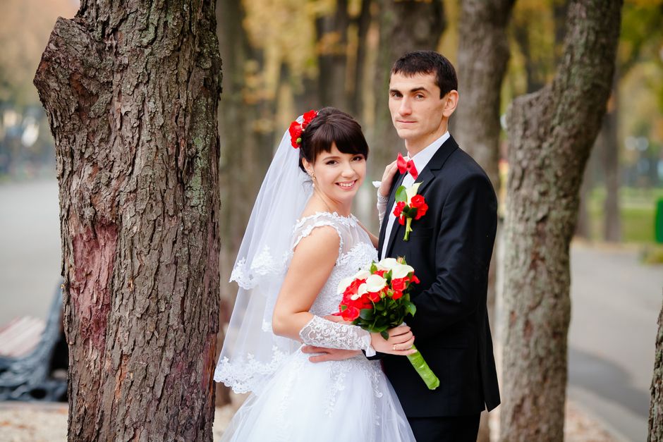 A bride and groom stand outdoors amidst tall, textured trees during daytime, with the bride holding a bouquet of red and white roses and dressed in a white lace wedding gown with a veil and floral hai