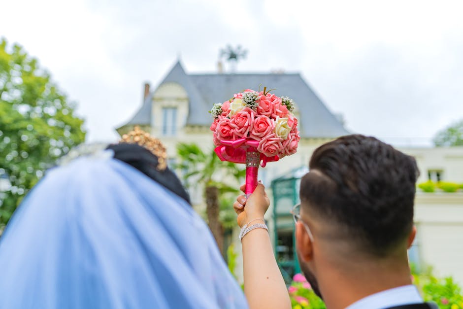 Close-up view of a wedding bouquet featuring tightly arranged pink and cream roses with green foliage accents, held by a person with a pink ribbon-wrapped handle. The bouquet is positioned in front of